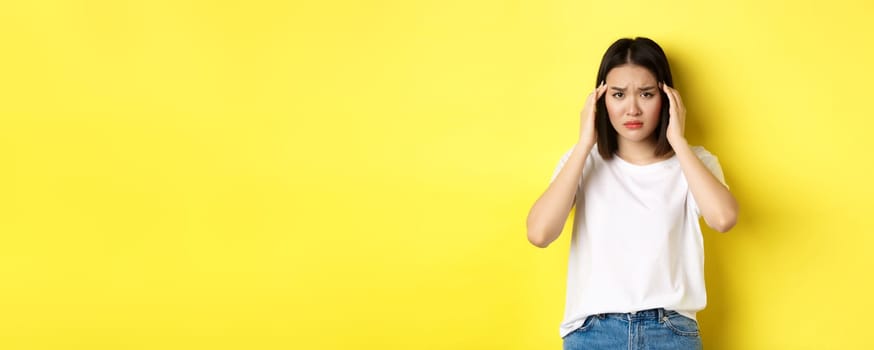 Sad asian girl touching head and frowning, feeling sick, having headache, standing in white t-shirt over yellow background.