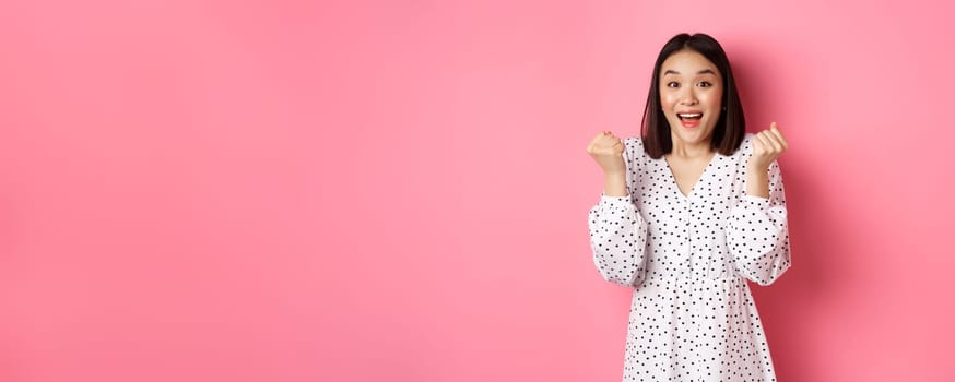 Image of beautiful asian woman winning, clench fists and saying yes with happy face, triumphing and celebrating victory, standing over pink background.