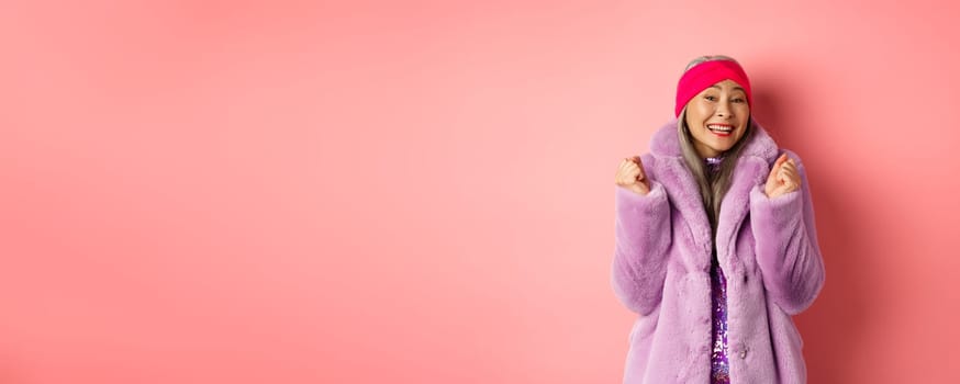 Cheerful asian lady in stylish funky coat, celebrating victory or success, saying yes and smiling happy, standing over pink background.