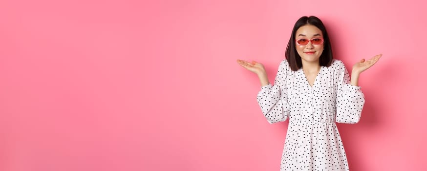 Cute asian woman tourist smiling at camera, shrugging clueless, dont know, wearing trendy sunglasses and white dress, standing against pink background.