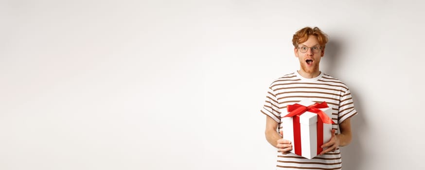 Valentines day and holidays concept. Surprised redhead boyfriend in glasses looking grateful at camera, receive big present in box, standing over white background.