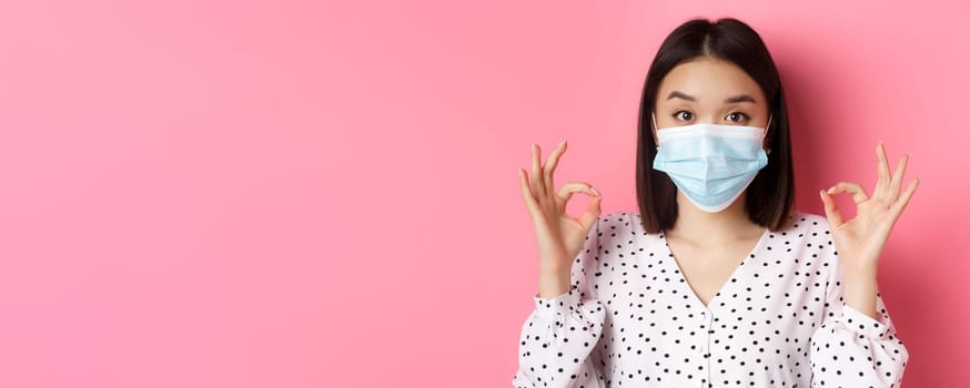 Covid-19, pandemic and lifestyle concept. Image of cute asian woman in face mask showing its okay gesture, make ok signs, have all under control, standing over pink background.