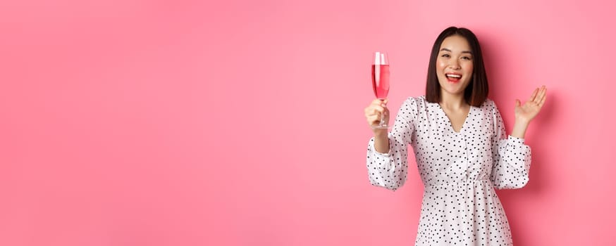 Happy asian woman celebrating, saying toast on party, raising glass of champagne and smiling, standing in dress over pink background.