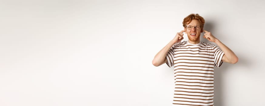 Annoyed redhead guy in glasses shutting ears and complaining loud noise, angry at noisy neighbours, standing over white background.