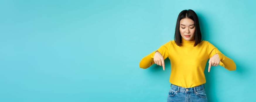 Shopping concept. Young teenage korean girl pointing and looking down at commercial, standing against blue background.