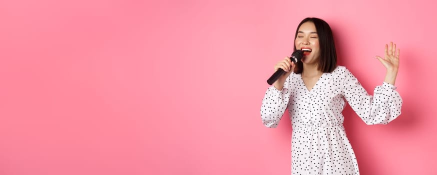 Happy asian woman dancing and singing in microphone, performing at karaoke, standing over pink background. Copy space