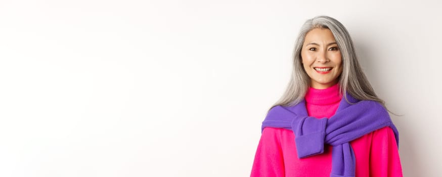 Close-up of beautiful asian senior woman in trendy pink sweater, smiling cheerful at camera, standing over white background.