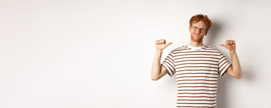 Confident young nerdy guy in glasses with red hair, pointing at himself and looking like professional, white background.