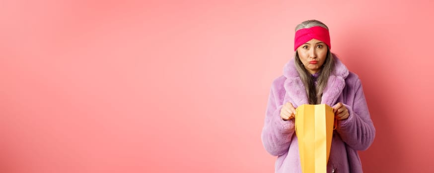 Upset senior asian woman open shopping bag and looking sad at camera, standing over pink background.