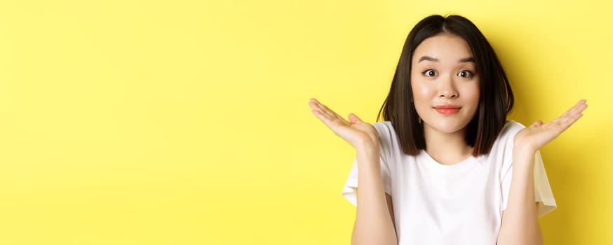 Silly mistake. Close up of cute asian girl saying sorry, shrugging shoulders and smiling with oops face expression, standing over yellow background.