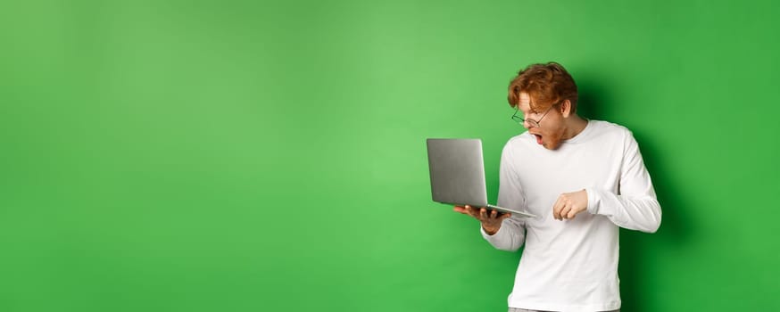 Image of young redhead man looking surprised at laptop screen, drop jaw and staring at display amazed, green background.