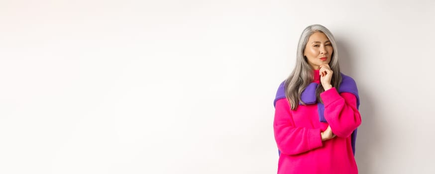 Image of thoughtful asian senior woman squinting suspicious at camera, having assumption, standing over white background and thinking.