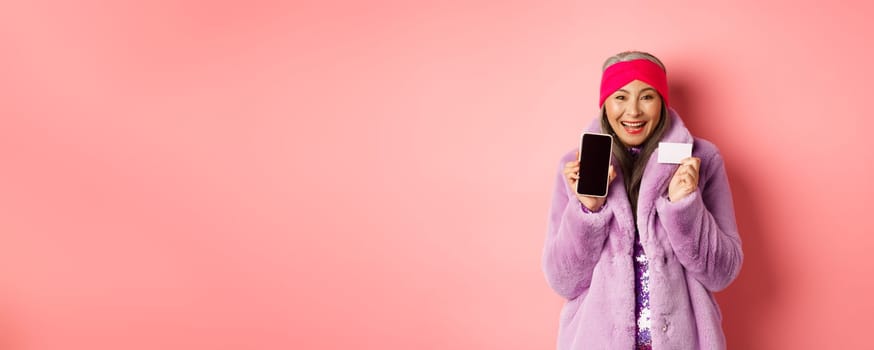 Online shopping and fashion concept. Beautiful asian mature woman showing plastic credit card and blank phone screen, smiling happy, pink background.