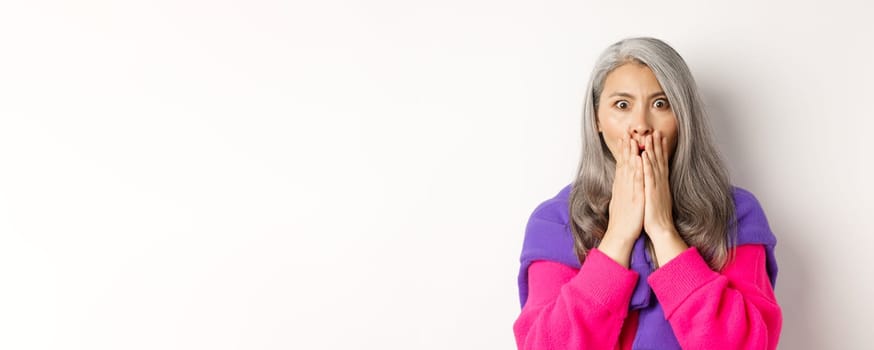 Close up of shocked asian old woman with grey hair, standing in stylish pink sweater, gasping startled and amazed, staring at camera, white background.