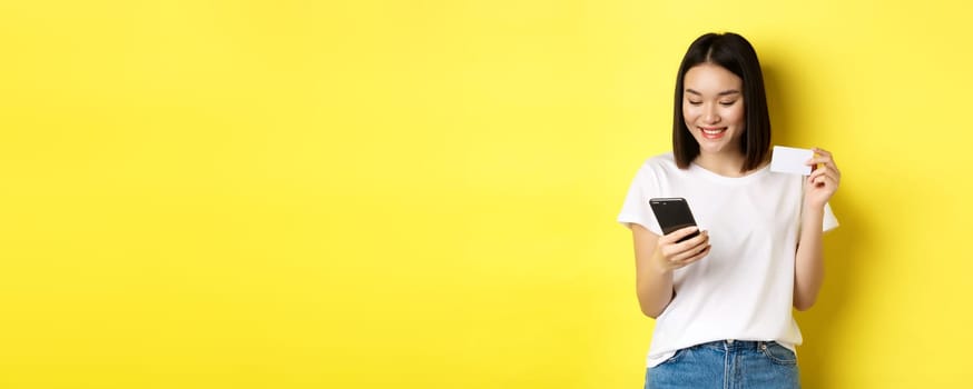 E-commerce and online shopping concept. Beautiful asian woman paying in internet, looking at smartphone screen and holding plastic credit card, yellow background.