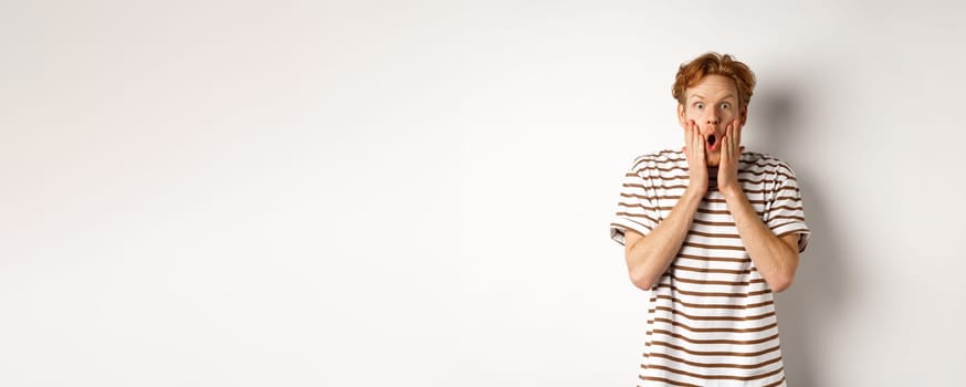 Impressed young man with ginger hair, gasping and staring shocked at camera, express complete disbelief and amazement, white background.