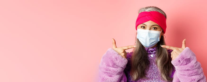 Covid-19, virus and fashion concept. Fashionable asian senior woman in trendy outfit pointing fingers at medical mask, follow social distancing rules, pink background.