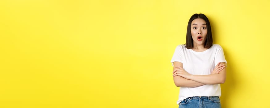 Portrait of surprised asian girl checking out promo, gasping amazed, staring at camera amazed, yellow background.