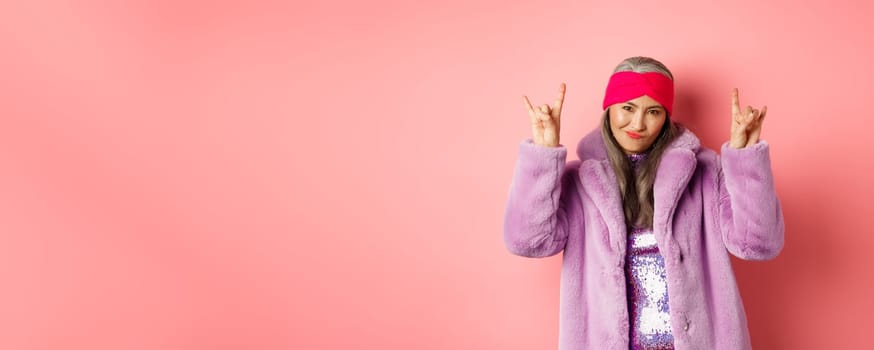 Cool asian mature woman in fashionable faux fur coat, showing rock-n-roll horns signs and smiling sassy at camera, having fun, standing over pink background.