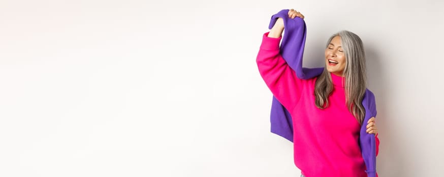 Carefree korean elderly woman in pink sweater, dancing with sweatshirt on shoulders and smiling, posing happy over white background.