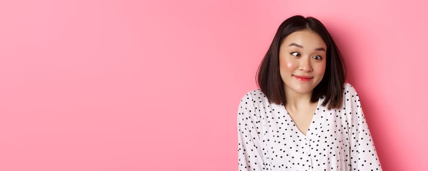 Beauty and lifestyle concept. Close-up of funny and cute asian woman squinting eyes and smiling, fool around at camera, standing over pink background.