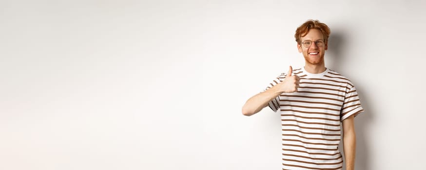Smiling young male student with red hair and bristle, showing thumb-up and looking satisfied, leave positive feedback, recommending promo, white background.