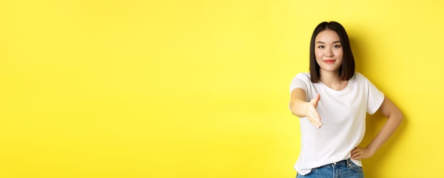 Confident asian woman in white t-shirt, stretch out hand for handshake and greeting gesture, saying hello, introduce herself, standing over yellow background.