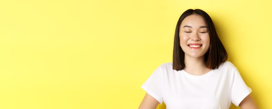 Close up of happy, romantic asian girl dreaming of something, close eyes and smiling delighted, standing over yellow background.