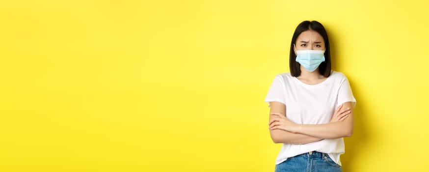 Covid, health care and pandemic concept. Asian woman in white t-shirt and medical mask cross arms on chest and looking sad and disappointed at camera.