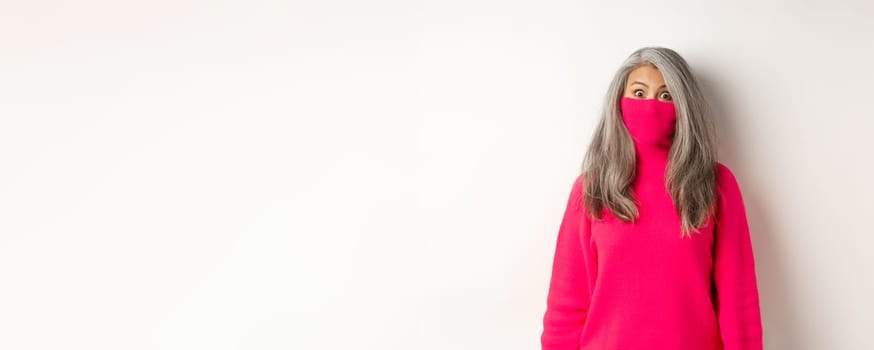 Funny and cute asian senior woman pull sweater collar on face and looking at camera, standing over white background.