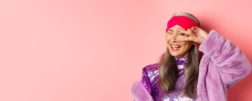Fashion. Close-up of fashionable asian senior woman smiling, showing victory sign over eye and looking happy at camera, standing over pink background.