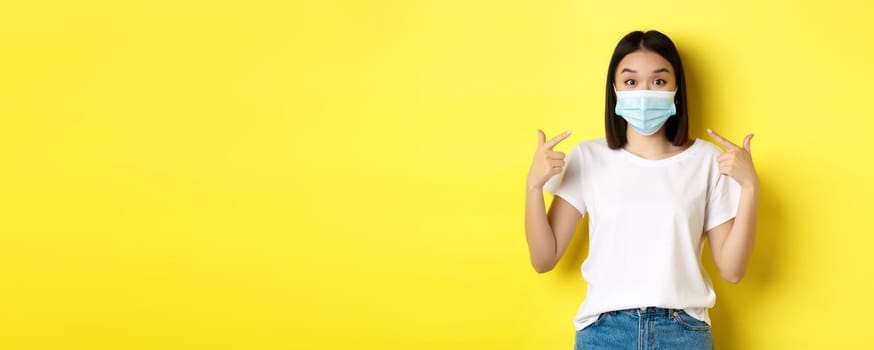 Covid-19, quarantine and social distancing concept. Young asian woman in white t-shirt, pointing at her medical mask from coronavirus, yellow background.