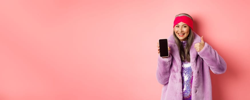 Online shopping and fashion concept. Funky asian senior woman showing blank mobile screen and thumbs-up, like and recommend internet promo, pink background.