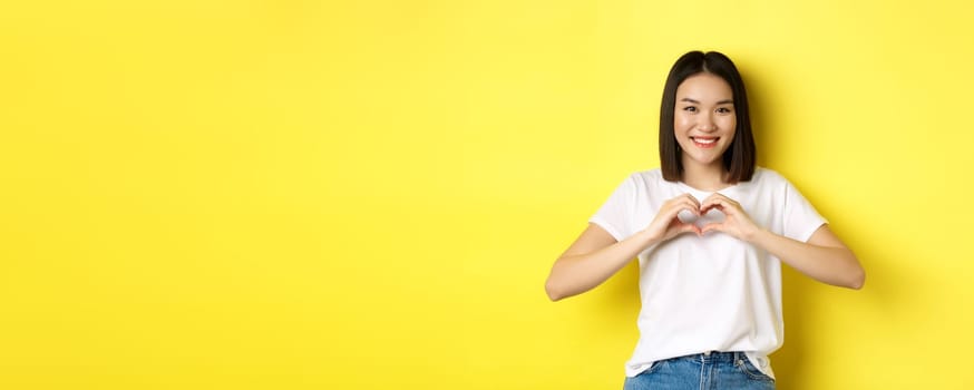 Beautiful asian woman showing I love you heart gesture, smiling at camera, standing against yellow background. Concept of valentines day and romance.