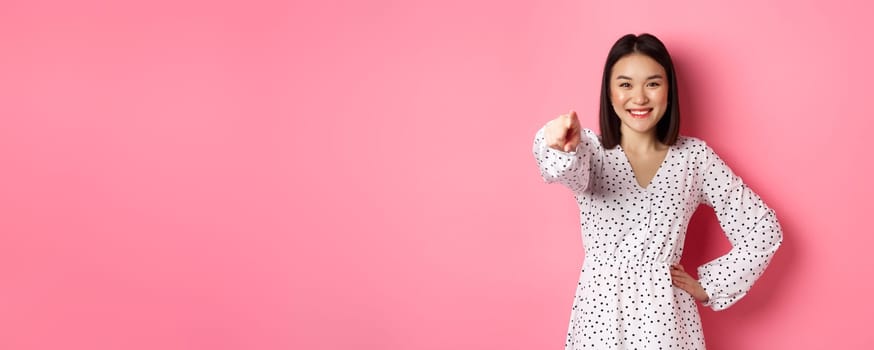 Confident asian woman in dress pointing at you, smiling and staring at camera, standing against pink background. Copy space