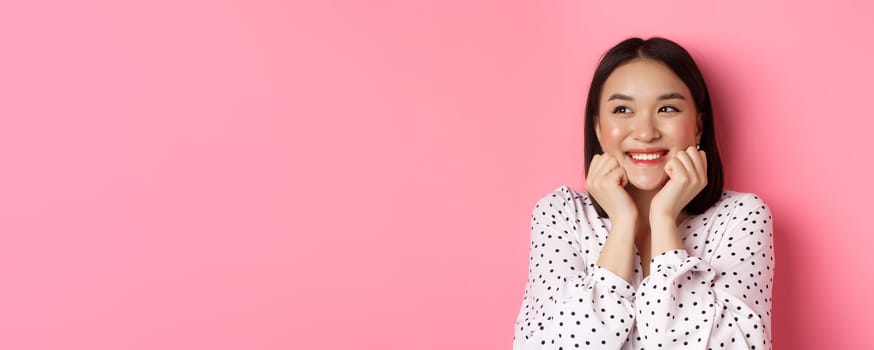 Beauty and skin care concept. Close-up of beautiful asian woman with clean face, blushing and looking dreamy left, admiring promo offer, standing over pink background.