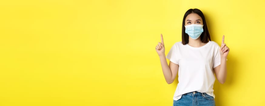 Covid-19, pandemic and social distancing concept. Young asian woman in white t-shirt and medical mask from coronavirus, looking and pointing fingers up, showing special offer.