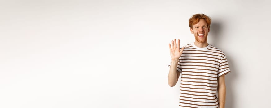 Friendly man with red hair and beard saying hi, waving hand and smiling, standing over white background.