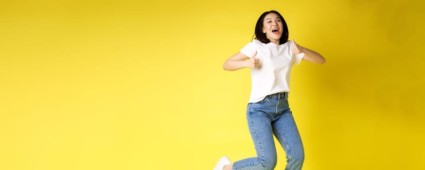 Full size of happy young asian woman jumping from joy, showing thumbs up in approval, posing over yellow background in jeans and casual white t-shirt.