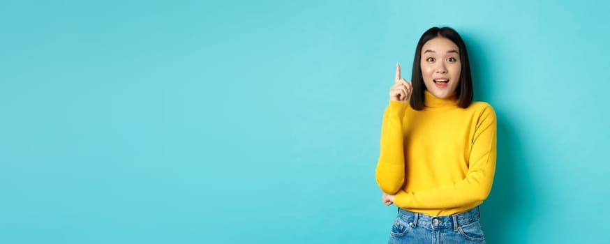 Excited asian woman with short dark hair, pitching an idea, raising finger in eureka gesture and smiling, standing over blue background.