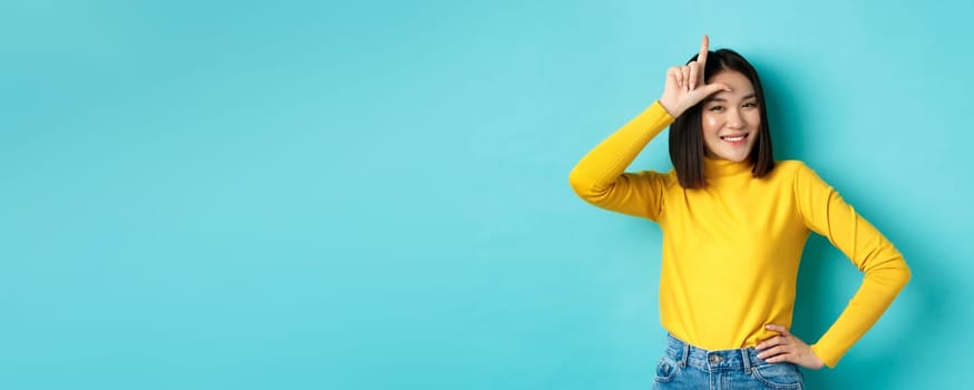 Sassy asian girl mocking lost team, showing loser sign on forehead and smiling pleased, being a winner, standing over blue background.