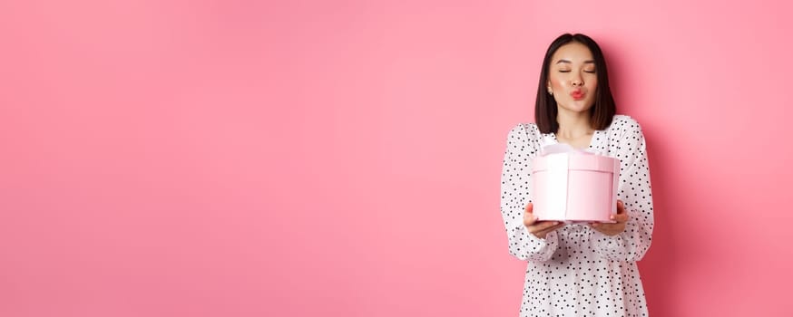 Cute asian girlfriend congratulate with valentines day, giving cute romantic gift in box, pucker lips for kiss, standing over pink background.