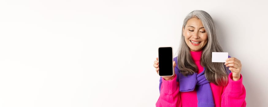 Online shopping. Closeup of fashionable old woman showing blank smartphone screen, looking pleased at plastic credit card, standing over white background.