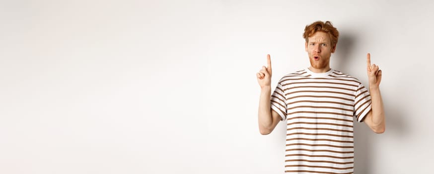 Confused young man with red curly hair pointing fingers up, staring shocked and puzzled, standing over white background.