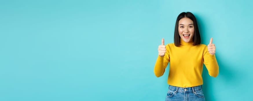 Beautiful asian woman praise good work, showing thumbs up gesture and smiling in approval, recommend product, standing satisfied over blue background.
