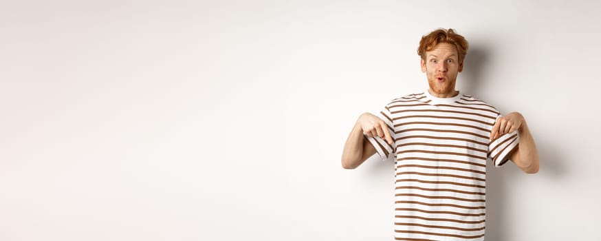 Image of funny redhead male student pointing fingers down, showing promo offer with excited smile, standing over white background.