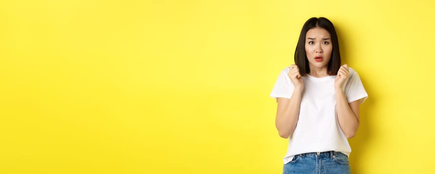 Beauty and fashion concept. Scared timid asian girl looking anxious, gasping startled and standing over yellow background.