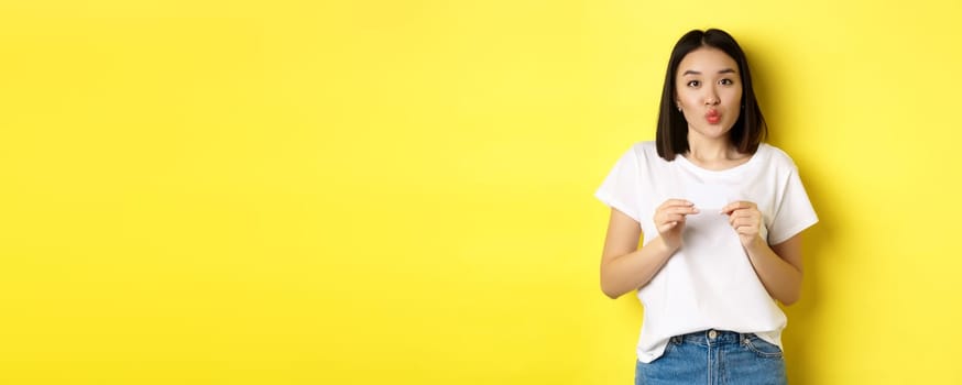Young asian woman in casual white t-shirt showing plastic credit card and pucker lips silly, going on shopping, yellow background.