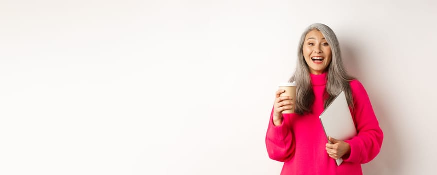 Happy asian senior female entrepreneur laughing, drinking coffee and holding laptop, standing in pink sweater over white background.