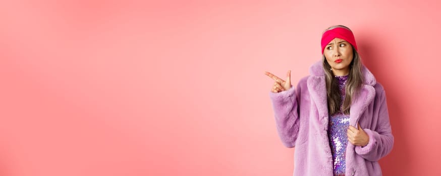 Fashion and shopping concept. Skeptical middle-aged asian woman in purple fur coat looking and pointing finger left with displeased, sad expression, standing over pink background.